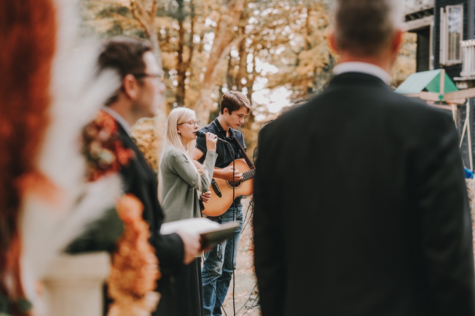 Sophie singing into a microphone and William playing guitar during an outdoor autumn wedding ceremony, guests visible in the foreground
