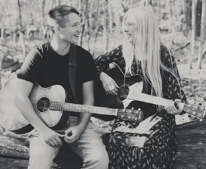 Black and white portrait of Sophie and William sitting on a fallen log outdoors, both playing acoustic guitars and laughing with each other