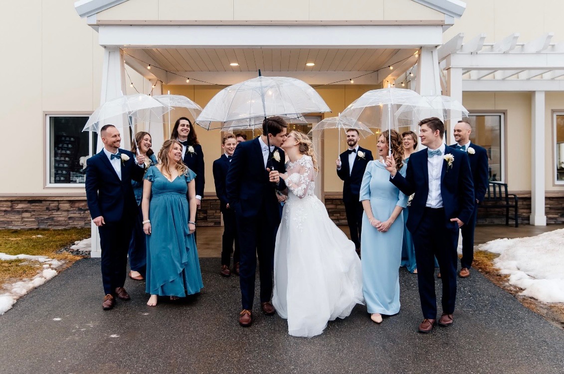 A joyful wedding party surrounding a bride and groom sharing a kiss, everyone holding clear bubble umbrellas on a winter wedding day