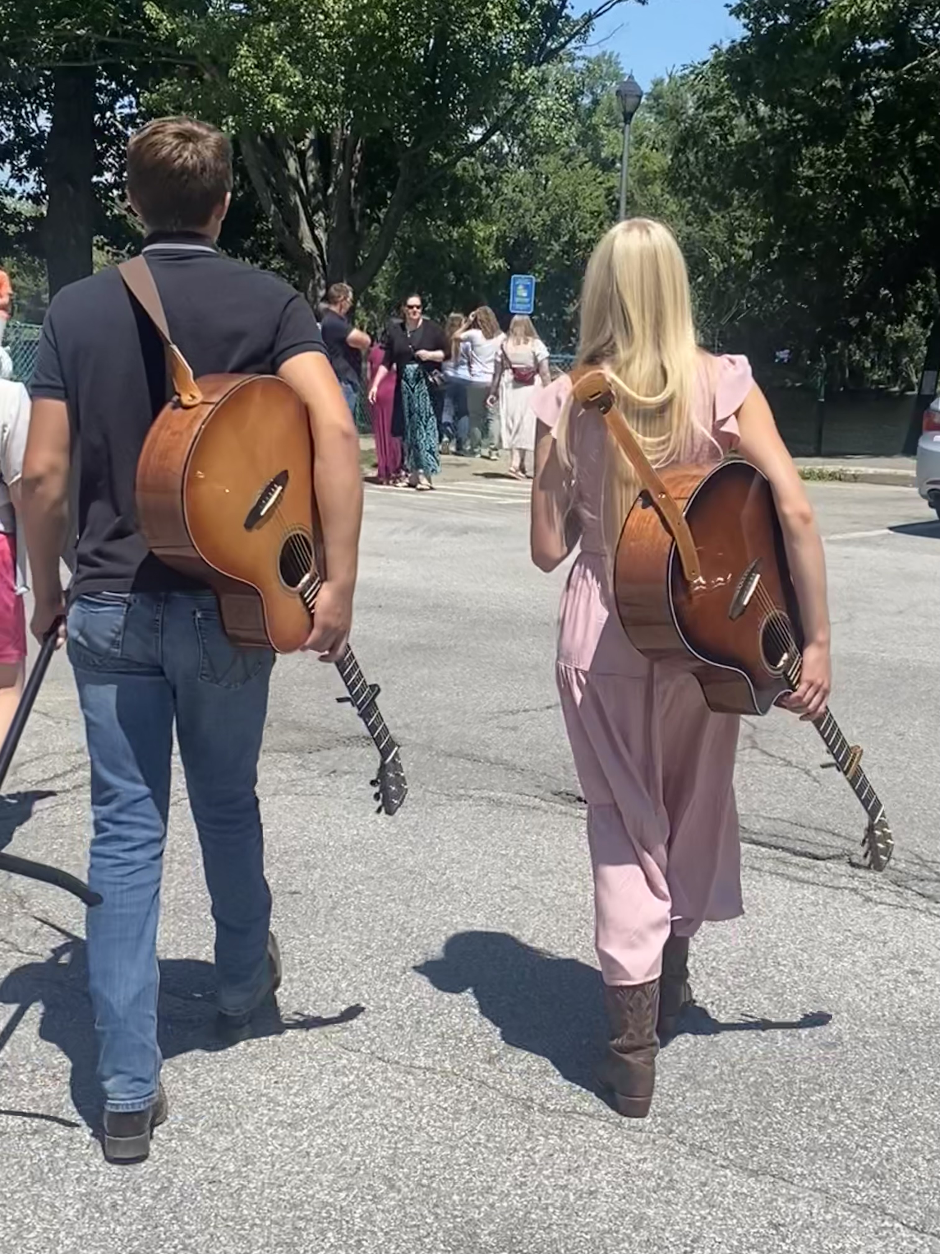 Sophie and William walking side by side, each with an acoustic guitar on their back, arriving to perform at a sunny outdoor event