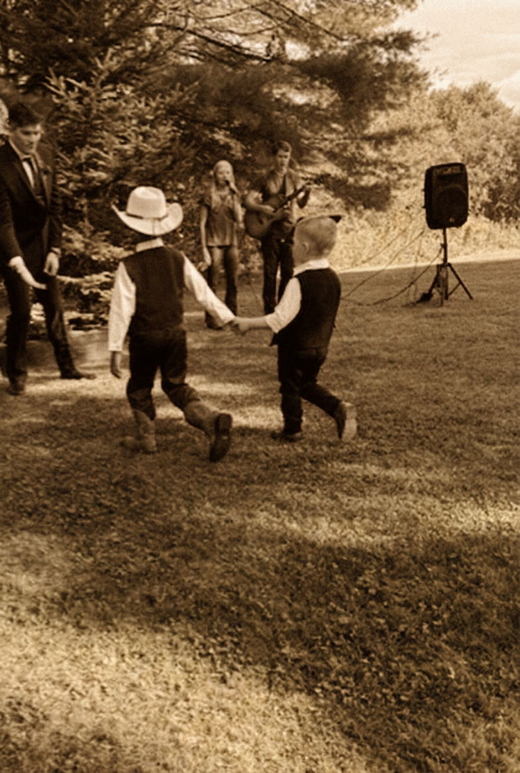 Two young boys in dress clothes spin and dance together on a grassy lawn while Sophie sings and William plays guitar at an outdoor wedding reception