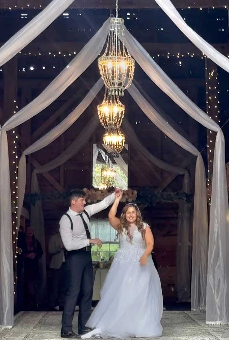 A bride beaming with joy as her groom spins her during their first dance beneath a glittering chandelier in a warmly lit barn wedding venue